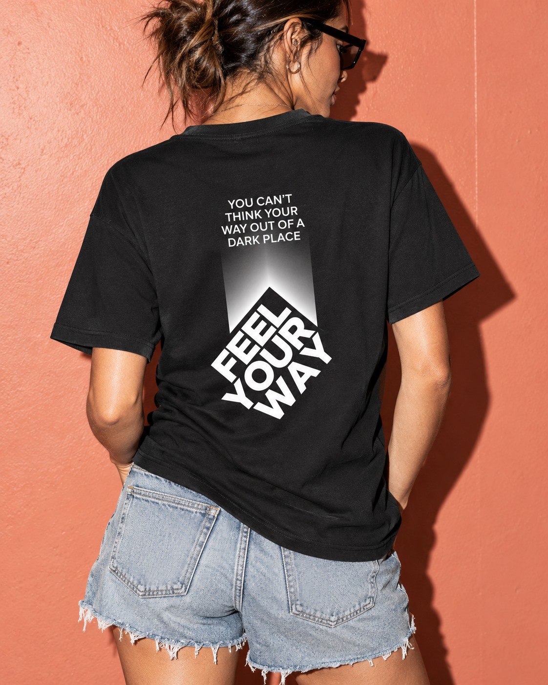 Woman wearing a black You Can’t Think Your Way, You Feel Your Way T-shirt against a coral wall.