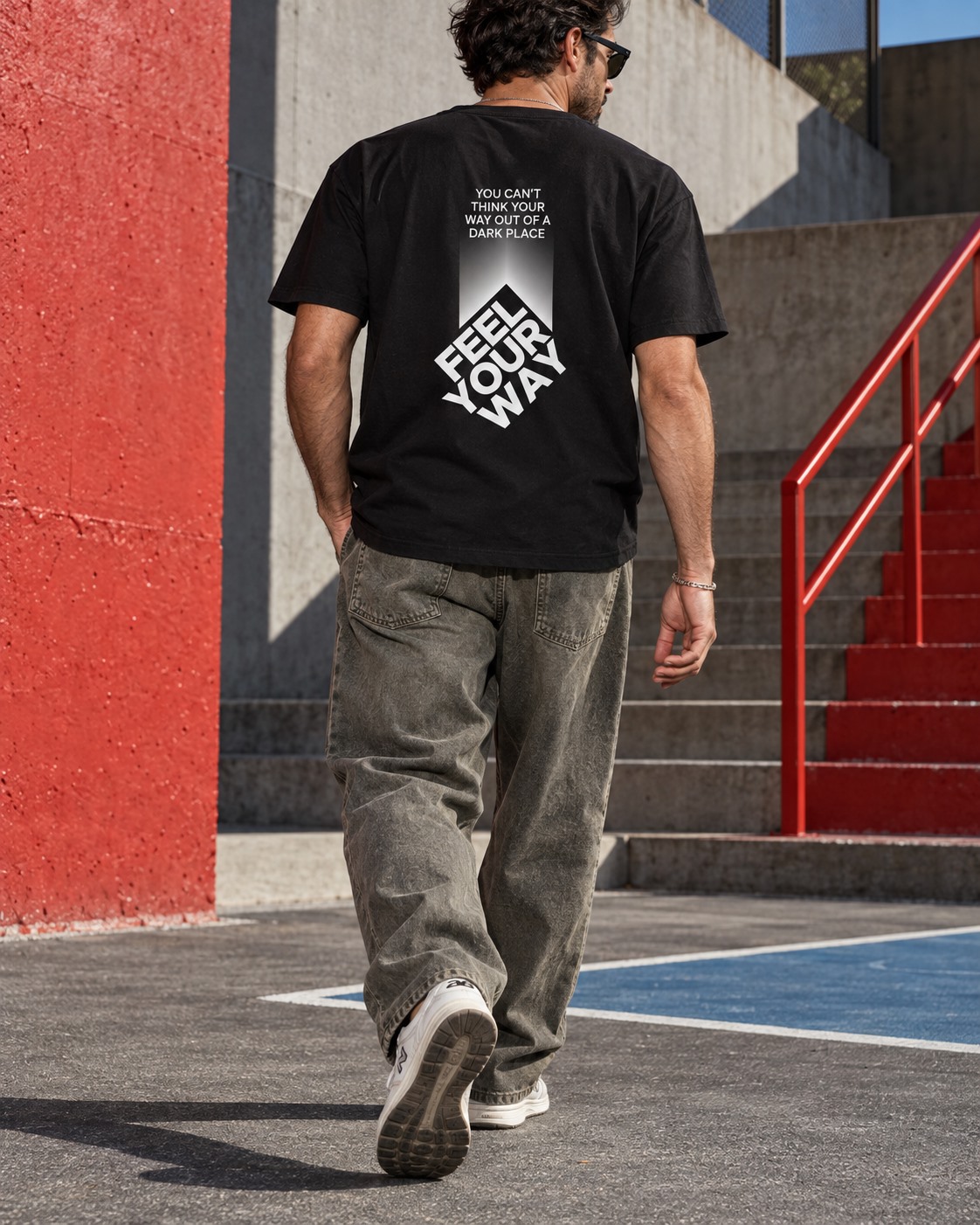 Man wearing a black You Can’t Think Your Way, You Feel Your Way T-shirt beside red stairs.
