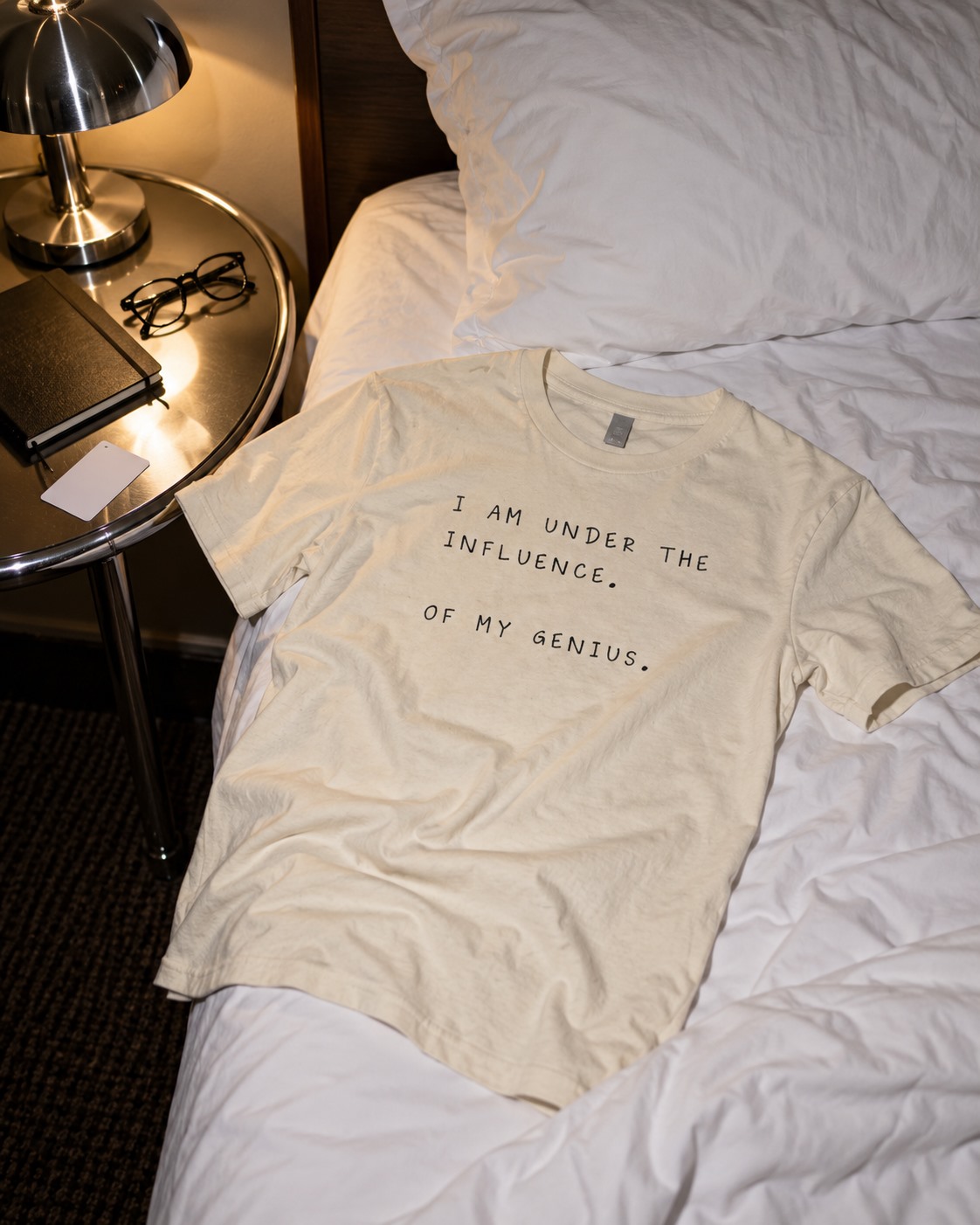 Cream Under the Influence of My Genius T-shirt laid on a bed.