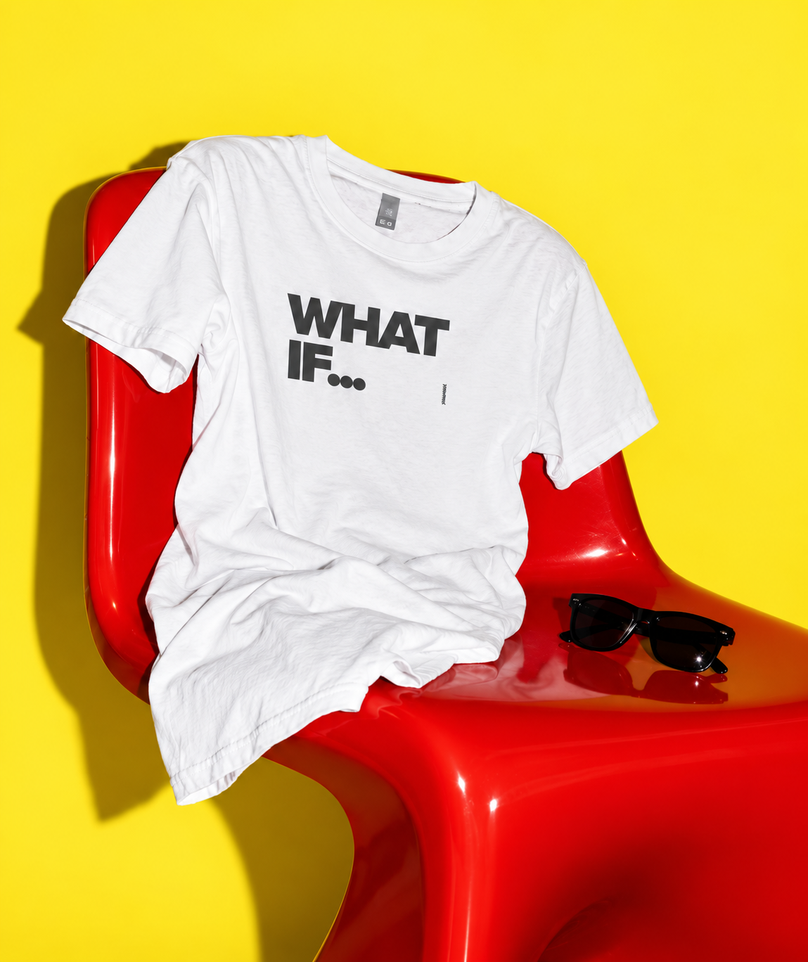 White What If T-shirt draped over a red chair against a vivid yellow background.